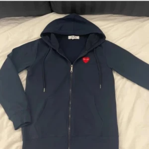 Mörkblå hoodie Comme des Garçons Play - Säljer en mörkblå hoodie från Comme des Garçons Play med dragkedja och huva. Tröjan har det ikoniska röda hjärtat med ögon broderat på bröstet. Långärmad och i mjuk bomull, perfekt för en chill och stilren look. Obs dragkedjan är trasig så det går ej att knäppa upp den hela vägen. Annaes helt ny