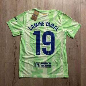Barcelona Lamine Yamal fotbollströja S - Säljer en officiell FC Barcelona fotbollströja med Lamine Yamal och nummer 19 på ryggen. Tröjan är ljusgrön med vita och gröna grafiska mönster, korta ärmar och rund hals. Märkt med Nike-logga, Spotify och UNHCR tryck samt FCB klubbmärke. Tillverkad i lätt polyester.