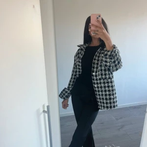 Rutig jacka/kappa  - Snygg overshirt i svart och vitt rutigt mönster med klassisk krage och knappar framtill. Jackan har en loose passform och är tillverkad i ett grovt, strukturerat material som ger en trendig look. Perfekt att slänga över en t-shirt eller stickad tröja.