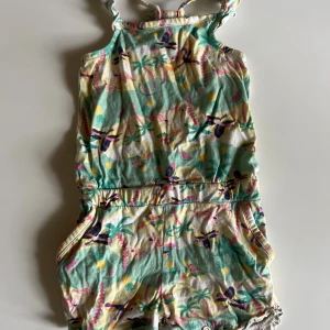 Färgglad playsuit med tropiskt mönster KappAhl - Superfin playsuit från KappAhl i storlek 98 med tropiskt mönster i grönt, gult, rosa och lila. Mjuk bomull, volangdetaljer på axelbanden och söt rosett baktill. Perfekt för varma sommardagar och lek utomhus.