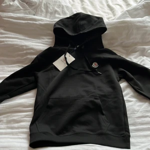 Svart hoodie från Moncler - Svart hoodie från Moncler med klassisk huva och dragsko, samt broderad Moncler-logga på bröstet. Tröjan har känguruficka framtill och ribbade muddar vid ärmslut och nederkant. Perfekt för en chill och stilren look.