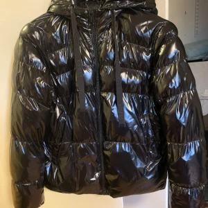Svart glansig pufferjacka från Guess - Svart pufferjacka från Guess med glansig finish och huva. Jackan har dragkedja framtill, breda kanaler och snörning i huvan. Snygg och modern look med Guess-logga på insidan. Snyggt sydd av skräddare på bild 4 då den gick sönder lite förra vintern, och även en liten fläck färg bortskavd vid andra fickan på sista bilden. Annars är den som i ny skick då jag gillar den mycket och har tagit hand om den väl. Nypris 2000. Hör av dig vid funderingar💕