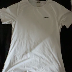 Vit t-shirt från Stronger - Vit t-shirt från Stronger med enkel design och liten svart logga på bröstet. Klassisk rund halsringning och korta ärmar. Tillverkad i mjukt och lätt polyestermaterial, perfekt för träning eller chill dagar.