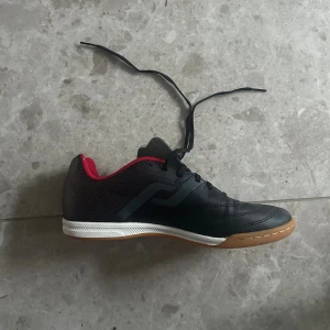 Svarta fotbollsskor Pro Touch  - Säljer ett par svarta fotbollsskor från Pro Touch med snygga röda detaljer och prickigt mönster på hälen. Skorna har snörning, platt sula och rund tå. Yttermaterialet är syntet och insidan är vadderad för extra komfort. Perfekt för inomhusspel.