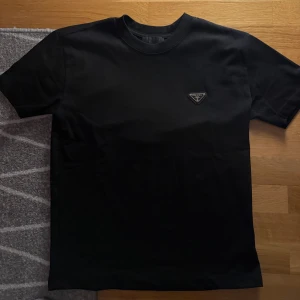 Svart t-shirt från Prada - Snygg svart t-shirt i Storlek S från Prada med klassisk rund hals och korta ärmar. På bröstet sitter Pradas ikoniska trekantiga logga i metall. T-shirten är gjord i mjuk bomull och har en clean, stilren look som passar till allt. Pris kan diskuteras vid snabb affär