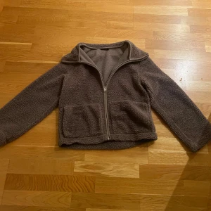 Brun teddyjacka med dragkedja - Mysig brun teddyjacka med hel dragkedja framtill och två stora fickor. Jackan har hög krage och är långärmad, perfekt för kyliga dagar. Den är i en loose passform och tillverkad i mjukt fleece-material.