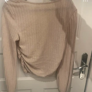 Beige ribbad långärmad tröja - Säljer en beige ribbad tröja med lång ärm och snygg rynkdetalj på ena sidan. Tröjan har en fyrkantig ringning och är i ett mjukt, stretchigt material som sitter skönt på kroppen. Perfekt att styla med jeans eller kjol.