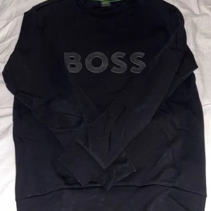 Svart långärmad tröja från BOSS - Säljer en svart långärmad tröja från BOSS med stor logga framtill. Tröjan har en klassisk rund halsringning och är tillverkad i mjukt bomullsmaterial. Perfekt för dig som gillar stilrena och enkla plagg med tydlig branding.