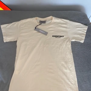 Beige Essentials Fear of God t-shirt - Snygg beige t-shirt från Essentials Fear of God i 100% bomull. Klassisk passform med rund hals och korta ärmar. Diskret logga på bröstet ger en clean och stilren vibe. Perfekt för dig som gillar minimalistisk streetwear.