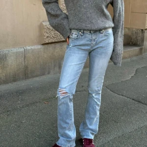 True Religion Becca Bootcut Jeans ljusblå - Ljusblå bootcut jeans från True Religion, modell Becca Mid Rise Bootcut. Snygga slitningar på ena benet, klassiska bakfickor med röd kontrastsöm och lock. Jeansen har medelhög midja och är tillverkade i jeansmaterial. Dem är i storlek W31 och passar mig som är 160cm och har storlek L! Dem är helt nya och nypris är 1299kr