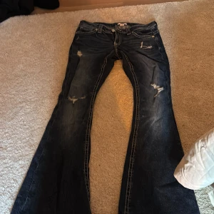 Mörkblå bootcut jeans Gina Tricot - Mörkblå bootcut jeans från Gina Tricot med slitna detaljer och snygga broderade bakfickor. Jeansen har kontrastsömmar och klassisk femficksdesign. Perfekta för dig som gillar en lite mer avslappnad och trendig look.