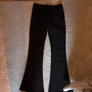 Svarta bootcut byxor med fickor - Snygga svarta bootcut byxor med hög midja och två bakfickor med lock och knapp. Byxorna har stängning med två knappar och dragkedja framtill. Materialet är slätt och har en lätt glansig finish som ger en stilren look. Helt nya aldrig använda och köpta för 400 kr