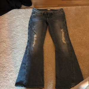 Bootcut jeans från Gina Tricot M - Bootcut jeans från Gina Tricot i mellanblå tvätt med slitna detaljer på låren och snygga bakfickor med lock. Jeansen har klassisk femficksdesign, låg midja och är tillverkade i jeansmaterial. Perfekta för dig som gillar en retro vibe och breda ben.