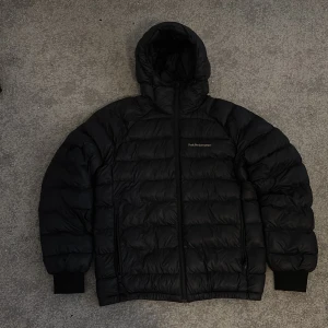 Svart vinterjacka från Peak Performance - Säljer en svart dunjacka från Peak Performance med huva och dragkedja. Nypris ~3000 kr, perfekt inför vintern. Jackan har ribbade muddar vid ärmsluten och en snygg remdetalj på huvan. Utmärkt för kalla dagar och mycket skön att bära. Hör gärna av dig om du har några frågor!