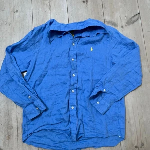 Blå linneskjorta från Polo Ralph Lauren - Snygg blå skjorta från Polo Ralph Lauren i luftig linnemix. Klassisk krage, långa ärmar och knappar framtill. På bröstet finns den ikoniska gula Polo-loggan som ger extra stilpoäng. Perfekt för dig som gillar en clean och fräsch look.