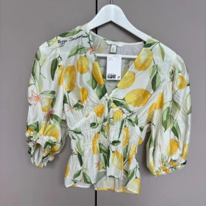 Blommig blus med citroner från H&M - Superfin blus från H&M med citron- och blommönster i gult, grönt och rosa på vit botten. Blusen har v-ringning, puffiga trekvartsärmar med resår och smockad midja som ger en snygg siluett. Perfekt för sommarens alla tillfällen! oanvänd