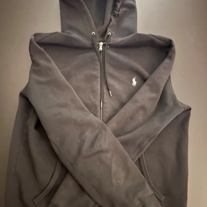 Svart hoodie från Polo Ralph Lauren - Svart hoodie från Polo Ralph Lauren med dragkedja framtill och klassisk broderad logga på bröstet. Tröjan har huva med snörning och fickor på sidorna. Perfekt för en chill och stilren look.