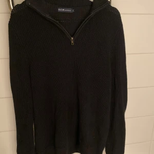 Svart stickad tröja med half zip S.C.W - Svart stickad tröja med halv dragkedja i halsen. Klassisk modell med lång ärm och ribbade muddar. Perfekt för kyliga dagar och enkel att matcha med jeans eller chinos. 