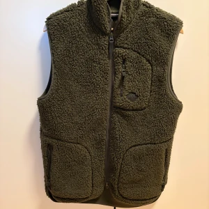 J.Lindeberg Fleeceväst - Säljer en olivgrön fleeceväst från J.Lindeberg med hög krage och hel dragkedja framtill. Västen har tre praktiska fickor med dragkedja och ett mjukt, fluffigt material som håller dig varm. Perfekt för lager-på-lager och chill stil. Nypris 1500kr