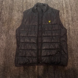 Svart dunväst från Lyle & Scott - Svart dunväst från Lyle & Scott. Västen har quiltad design och dragkedja framtill. Perfekt att slänga över en hoodie eller t-shirt för extra värme. Snygg, stilren och enkel att matcha med det mesta.