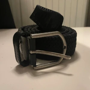 Svart flätat bälte med silverfärgat spänne - Snyggt svart flätat bälte med ett robust silverfärgat metallspänne. Bältet har detaljer i skinn vid ändarna och passar perfekt till jeans eller chinos. Enkel och stilren design som funkar till många outfits.