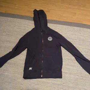 Mörkblå hoodie från Hackett London - Snygg mörkblå hoodie från Hackett London med dragkedja och huva. Tröjan har ett broderat märke på bröstet och två fickor framtill. Perfekt för dig som gillar klassisk stil med sportig touch.