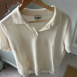 Vit pikétröja i kashmir från Soft Goat - Superskön vit pikétröja från Soft Goat i mjuk kashmir. Tröjan har klassisk krage, korta ärmar och två knappar framtill. Perfekt för dig som gillar stilrena och lyxiga plagg med en chill vibe.