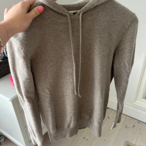 Beige hoodie i kashmir från Soft Goat - Mjuk och lyxig beige hoodie från Soft Goat i kashmir. Tröjan har huva med snörning, ribbade muddar och lång ärm. Perfekt för dig som gillar chill och stilren look. Snygg att matcha med jeans eller mjukisbyxor.
