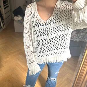 Vit stickad tröja med fransar - Säljer en vit stickad tröja med snyggt hålmönster och fransar vid ärmslut och nederkant. Tröjan har snörning vid halsen och lång ärm, perfekt för dig som gillar boho-vibe och vill ha något luftigt och coolt.