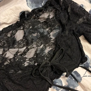 Svart spetsig långärmad korsettopp - Säljer en svart långärmad korsettopp i spets med blommigt mönster. Toppen har snörning framtill och är croppad med transparenta ärmar och överdel. Perfekt för dig som vill sticka ut med en edgy och trendig look.