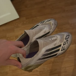 Adidas F50  - Säljer ett par Adidas F50 fotbollsskor i vitt med svarta och guldiga detaljer. Skorna har en strömlinjeformad design med svarta ränder på sidorna och guld på hälen och sulan. Ovandelen är i syntetmaterial och sulan har fasta dobbar för gräsplan.