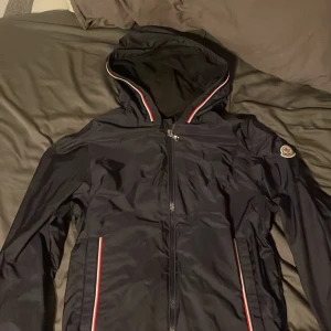 Mörkblå vindjacka från Moncler med huva - Mörkblå vindjacka från Moncler med huva och snygga rödvita detaljer längs dragkedjan och fickorna. Jackan har Moncler-logga på ärmen och hel dragkedja framtill. Perfekt för dig som gillar stilrena och sportiga plagg med exklusiv känsla. Köpt i Rom därav italiensk text på insidan. NFC Scan inuti jackan vid qr koden. Moncler Granduc. Og pris 6000 kr.