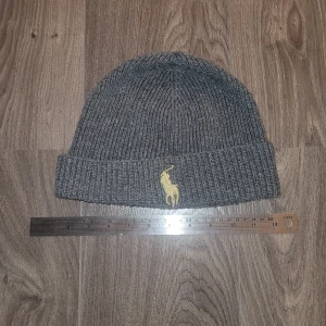 Grå ribbstickad mössa Polo Ralph Lauren - Snygg grå ribbstickad mössa från Polo Ralph Lauren med klassisk gul broderad logga framtill. Mössan har uppvikt kant och är tillverkad i mjukt material som håller dig varm under kyliga dagar. Perfekt accessoar för en clean och stilren look.