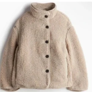 Beige teddyjacka med stora knappar - Mysig beige teddyjacka med hög krage och stora svarta knappar framtill. Jackan har en loose passform och är tillverkad i fluffigt fleece-material som håller dig varm. Perfekt för kyliga dagar och enkel att styla med olika outfits.