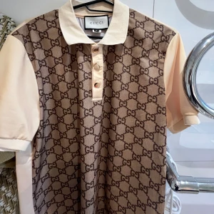 Beige GG-mönstrad skjorta från Gucci - Snygg beige skjorta från Gucci med klassiskt GG-mönster i brunt på framsidan. Skjortan har korta ärmar, krage och tre knappar framtill. Materialet känns mjukt och lyxigt, perfekt för dig som gillar designerplagg med tydlig logga.