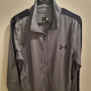 Grå jacka från Under Armour - Snygg grå jacka från Under Armour med svarta detaljer på axlarna och diskret logga på bröstet. Fast pris. Inga byten. Se gärna mina andra annonser.