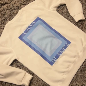 Vit sweatshirt från Casablanca M - Säljer en vit sweatshirt från Casablanca med blått grafiskt tryck både fram och bak. Tröjan har rund hals, långa ärmar och ribbade muddar. Perfekt för dig som gillar stilren design med en twist. Materialet är mjukt och skönt, troligtvis bomull.