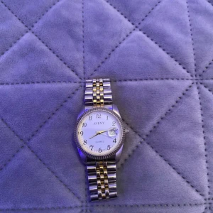 Aveny silver/guld armbandsur - Snygg klocka från Aveny med klassisk rund urtavla i vitt och tydliga svarta siffror. Armbandet är i silver och guldtonat metall med länkdesign. Klockan har datumvisning och guldfärgade visare som ger en stilren vibe. Perfekt accessoar för dig som gillar detaljer.