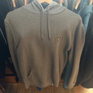 Grå hoodie från Lyle & Scott - Snygg grå hoodie från Lyle & Scott med klassisk gul logga på bröstet. Tröjan har huva med snörning och en stor magficka framtill. Perfekt för chill dagar och enkel att matcha med jeans eller joggers.