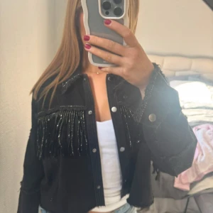 Svart jeansjacka med fransar från zara - Säljer en svart jeansjacka med coola fransdetaljer och pärlor både fram och bak. Jackan är croppad med tryckknappar och har en loose passform. Perfekt att styla med jeans eller kjol för en trendig look.
