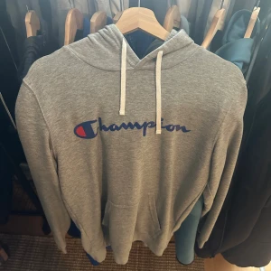 Grå Champion hoodie med snörning - Grå hoodie från Champion med klassisk logga i blått och rött på bröstet. Tröjan har vit snörning i huvan, känguruficka framtill och lång ärm. Perfekt för chill dagar och streetwear-stil.