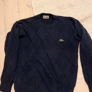 Mörkblå stickad tröja från Lacoste - Snygg mörkblå stickad tröja från Lacoste med klassisk krokodillogga på bröstet. Tröjan har ett diskret rutmönster i stickningen och rund hals. Perfekt för dig som gillar stilrena och tidlösa plagg med lite extra detalj.
