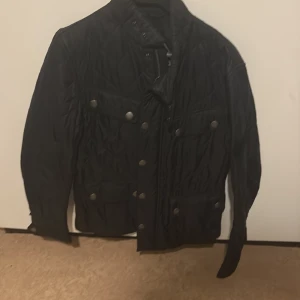 Svart quiltad jacka från Barbour S - Quiltad svart jacka från Barbour med coola tryckknappar och flera fickor framtill. Jackan har hög krage och klassisk bikerstil, perfekt för dig som gillar en stilren och tidlös look. Fodret har ett unikt tryck med motorcykelmotiv.