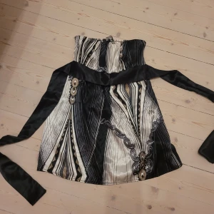 Svartvit topp från Lipsy London - Supersnygg topp från Lipsy London i svart, vitt och beige med grafiskt mönster. Toppen är axelbandslös med smock upptill och har ett brett satinband under bysten som kan knytas till rosett. Materialet är glansigt och känns lyxigt. Aldrig använd få den är för liten tyvärr... passar xxs-s med mindre byst🩷 så så fin!! Köpt för 1500