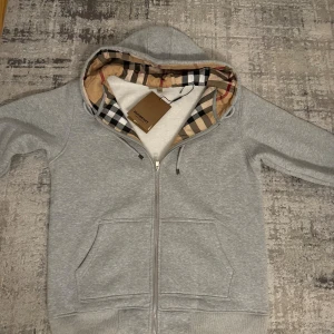 Grå Burberry zip hoodie med dragkedja - Snygg grå hoodie från Burberry med klassiskt rutigt foder i huvan och broderad logga på bröstet. Tröjan har hel dragkedja, snörning i huvan och stora fickor framtill. Perfekt för dig som gillar stilrena och lyxiga streetwear-plagg.