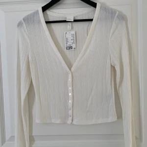 Vit ribbad blus från H&M - Säljer en fräsch vit blus från H&M i storlek S. Den har ribbad struktur, v-ringning och knappar framtill. Långärmad och croppad passform som ger en clean och modern look. Perfekt att styla med jeans eller kjol för en chill vibe.