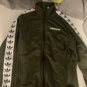 Grön Adidas track jacket XS - Snygg grön Adidas track jacket med vita ränder och svarta trefoil-loggor längs ärmarna. Jackan har hög krage, hel dragkedja och Adidas-logga på bröstet. Perfekt för en sportig och trendig look.