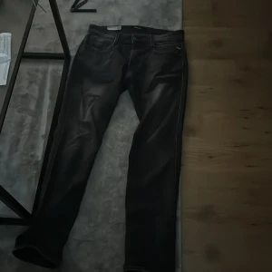 Grå Replay jeans med svart patch - Säljer ett par grå jeans från Replay med klassisk femficksdesign och svart läderpatch i midjan. Jeansen har raka ben och tydliga sömmar som ger en snygg kontrast. Perfekta för dig som gillar en stilren och avslappnad look.