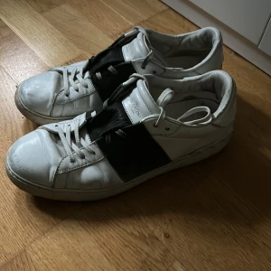 Valentino Garavani sneakers svart/vit - Snygga sneakers från Valentino Garavani i vitt skinn med en bred svart läderdetalj över mitten. Klassisk rund tå, vita snören och diskret logga på plösen. Perfekta för dig som gillar stilrena och exklusiva sneakers med en twist.