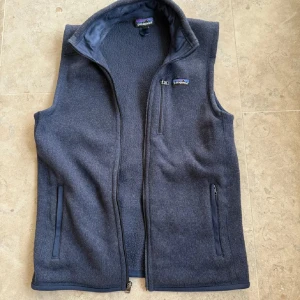 Mörkblå fleeceväst från Patagonia - Mysig mörkblå fleeceväst från Patagonia med hög krage och tre praktiska fickor med dragkedja. Perfekt att slänga på sig när det är lite kyligt ute. Västen har en stilren design och känns riktigt skön mot kroppen. Finns inte att köpa längre ny